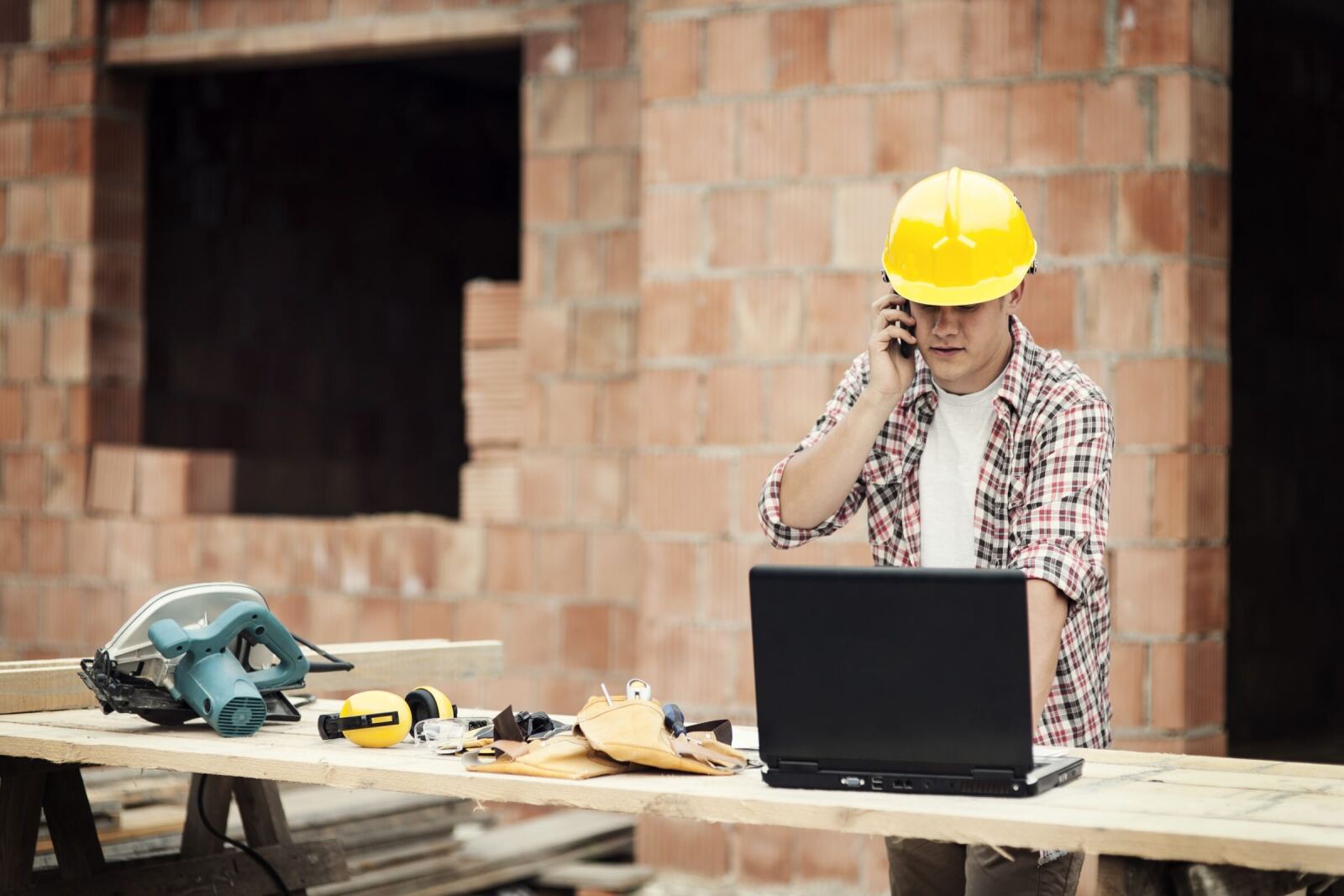 A carpenter on the phone at a site bench, laughing, with drawings and a laptop in front of him