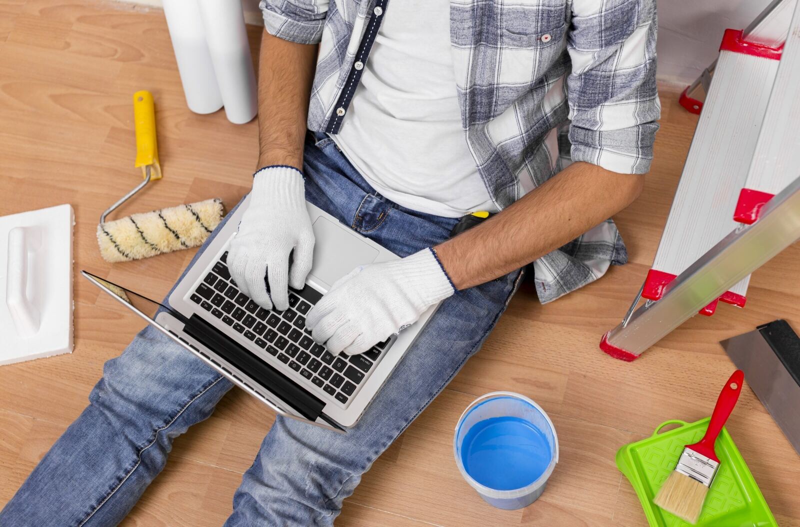 A tradesman sitting on a new timber floor with a laptop, roller and tools beside him, pricing a job