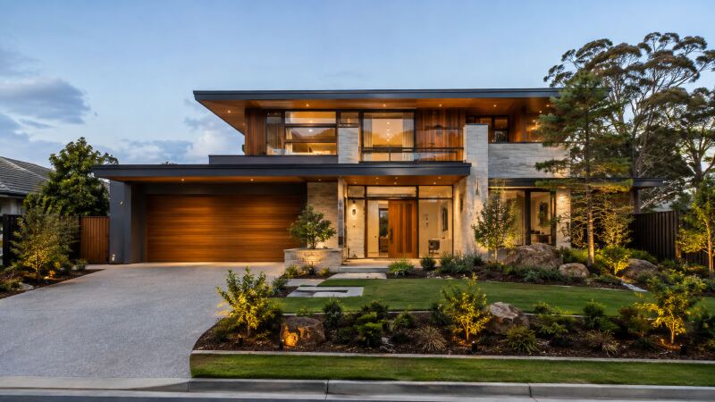 A completed two-storey home at dusk, lit from within, with timber and stone to the street